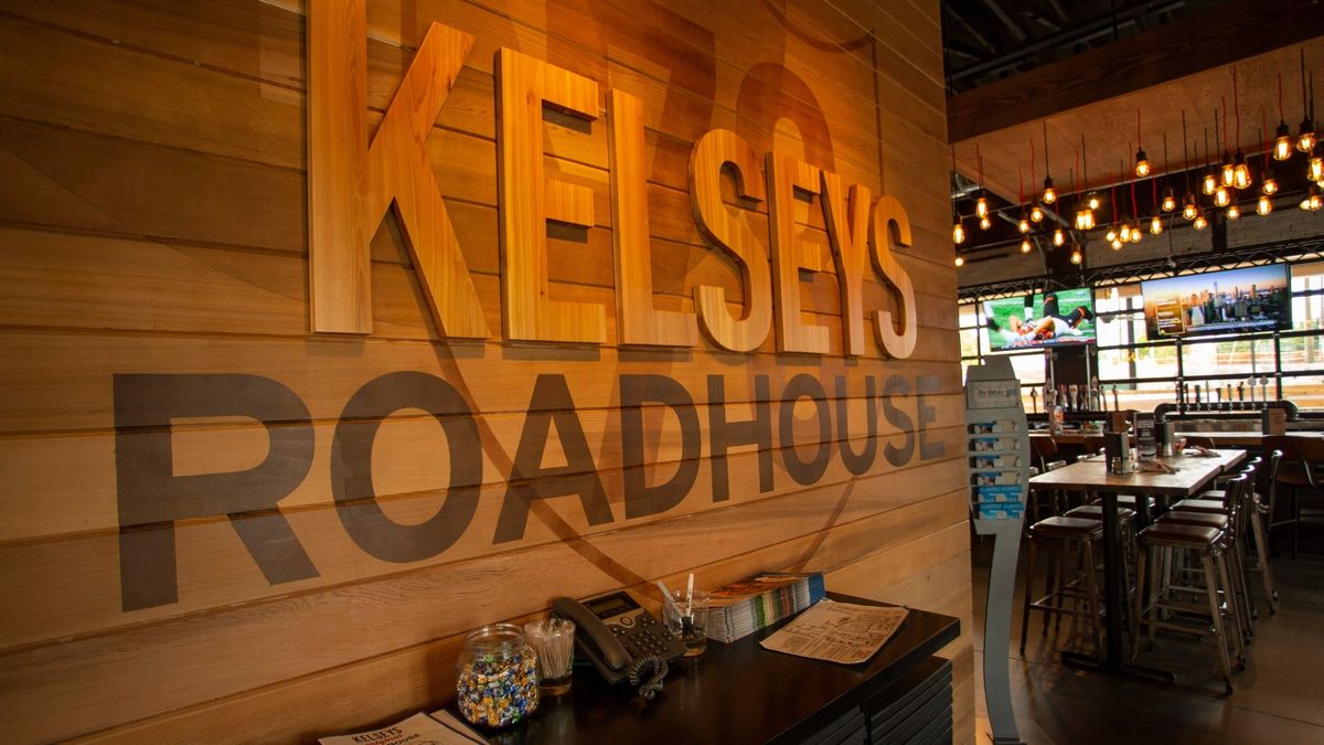 Inside Kelseys Original Roadhouse, with the Kelseys Roadhouse name on a wood wall and a bar under string lights