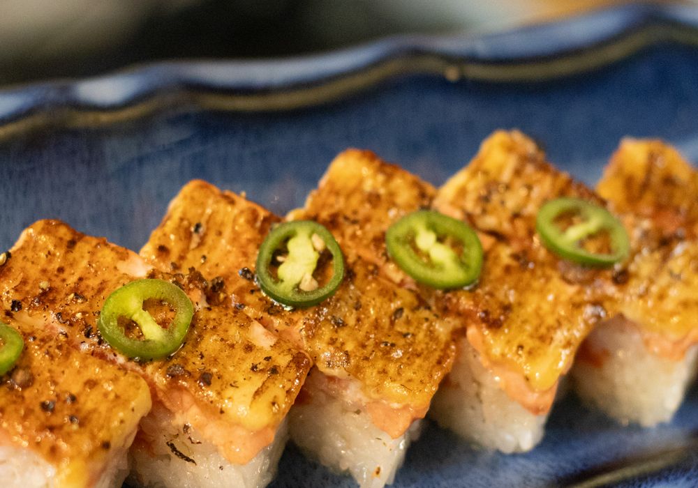 A row of pressed oshi sushi pieces, seared on top and each topped with a slice of jalape&ntilde;o, on a blue plate at Nare Japanese Restaurant