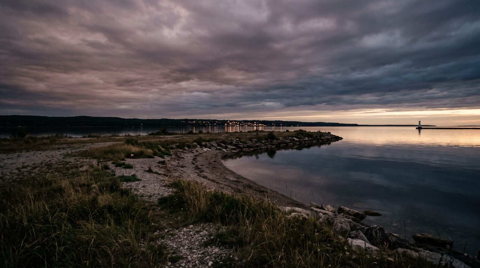 A former industrial waterfront point on Georgian Bay at twilight, open shoreline meeting still water with distant town lights and a slim lighthouse pier